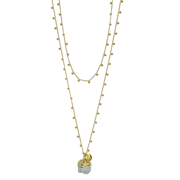 The Khione Necklace by Aria V is a layered piece featuring 16k gold plated chains. One chain is adorned with small, evenly spaced bead-like decorations, while the lower chain holds a large, irregularly shaped clear quartz pendant and a smaller, round, textured charm. The necklace has a delicate and elegant design.