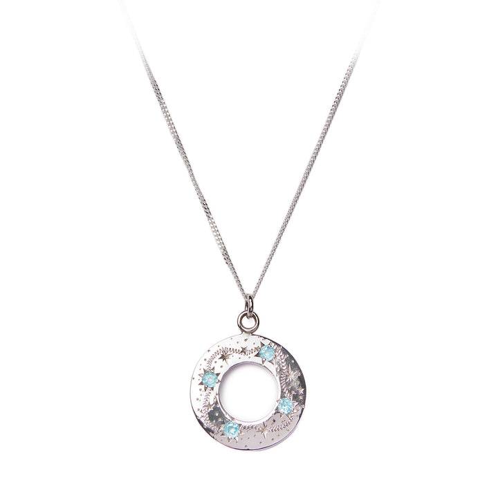 Introducing the "Something Blue" Shooting Star Pendant from Cosmic Boulevard. This exquisite necklace features a circular pendant intricately engraved with patterns and adorned with blue topaz gemstones, reminiscent of shooting stars. The delicate chain enhances the subtle elegance of this silver piece, making it a perfect accessory for sky lovers.