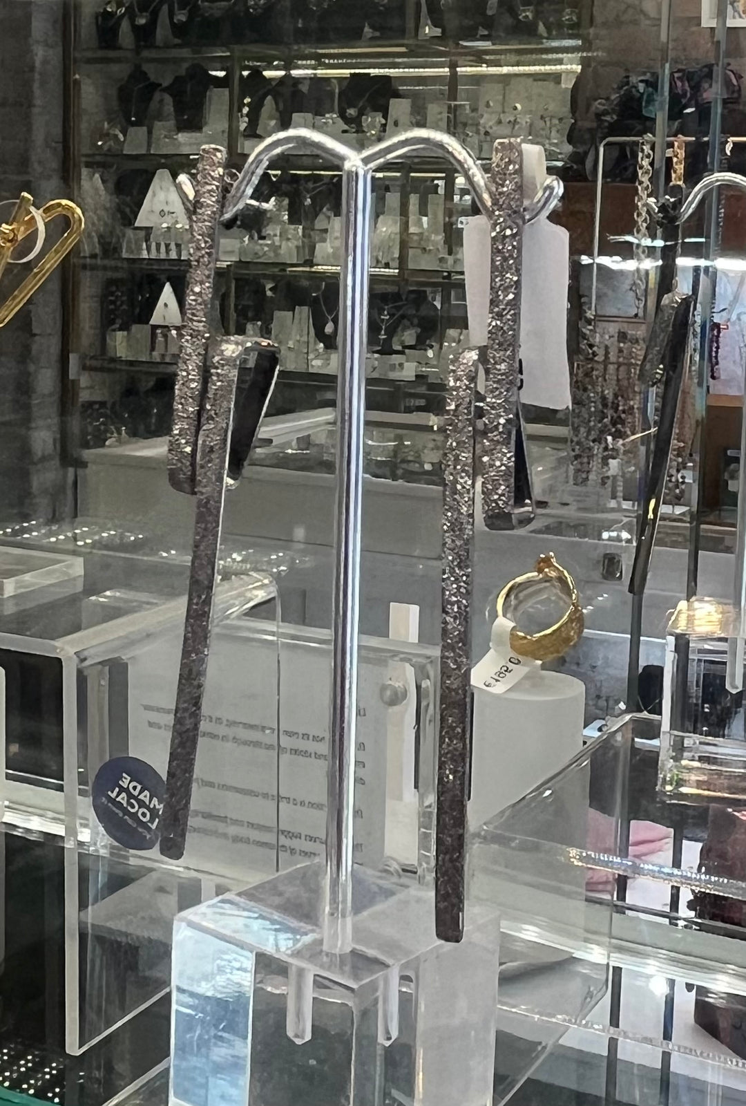 A display of modern, metallic Lysithea Black Rhodium Silver Earrings handcrafted by Theodora D on a transparent acrylic stand, set against a backdrop of various jewelry items in a glass showcase. The elongated, textured silver earrings with black rhodium plating hang gracefully from the stand.