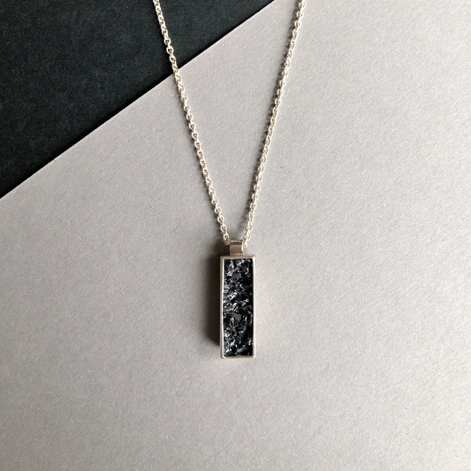 An Ingot Pendant by Angela O'Keefe is shown. The pendant features a rough black stone, possibly crushed silicon carbide, encased in an intricate silver frame. The necklace chain is visible, set against a background composed of contrasting black and light gray areas, evoking a gothic fairy tale aesthetic.