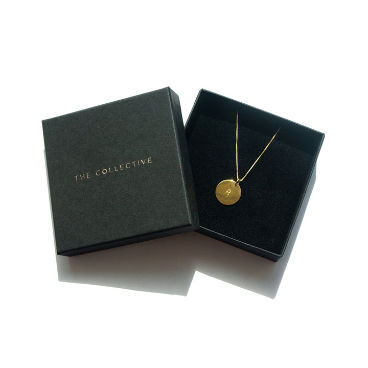 A black box with "Personalised Charms" written in gold text contains a Personalised Large Disc Charm in Gold. The pendant, adorned with a hand stamped charm bearing an engraved letter "R," hangs on an adjustable oval belcher chain. The open box displays the beautiful necklace against a black cushion.