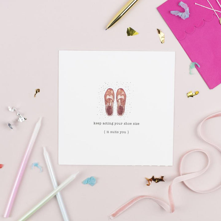 A beautifully textured card with an original illustration of two brown shoes and the text "KEEP ACTING YOUR SHOE SIZE { it suits you }" by Pickled Pompom. The card is surrounded by scattered colorful confetti, ribbon, and stationery items, including a pink Kraft envelope, white candles, and a gold pen on a pink surface.