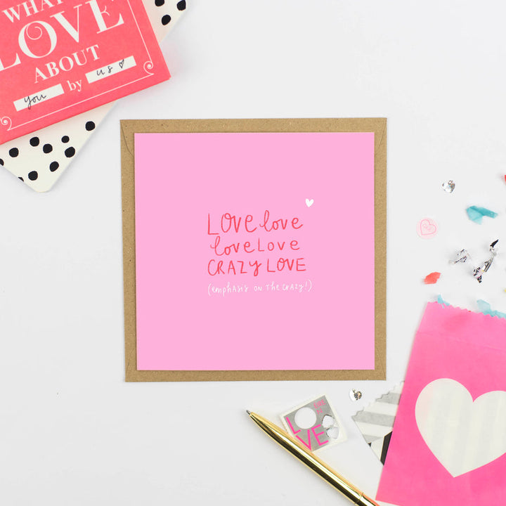 A pink greeting card with a brown border is on a white surface, accompanied by a pink kraft envelope, a red and white notebook, and some decorative items. The card, with an original illustration, reads "LOVE LOVE LOVE LOVE CRAZY LOVE (emphasis on the crazy)." This beautiful card is from Pickled Pompom's collection.