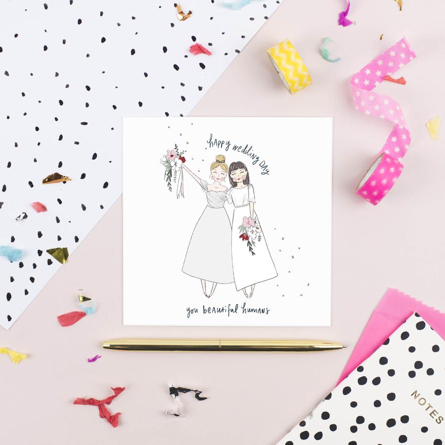 A textured card featuring an original illustration of two brides in white dresses, holding bouquets. The HAPPY WEDDING DAY GALS by Pickled Pompom reads "Happy wedding day, you beautiful humans" and is surrounded by confetti, ribbon, a golden pen, and polka-dotted paper and envelopes.