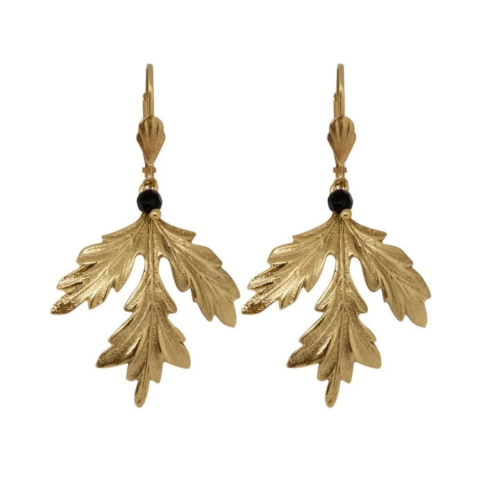 The Ella Maple Leaf Earrings Gold, a pair of gold-toned, nickel-free earrings designed in the shape of detailed, ornate leaves, each featuring a small, round black bead at the top where the leaf design hangs from the hook. Crafted by Lotta et Djossou from polished gold-plated brass, these earrings highlight intricate leaf patterns.