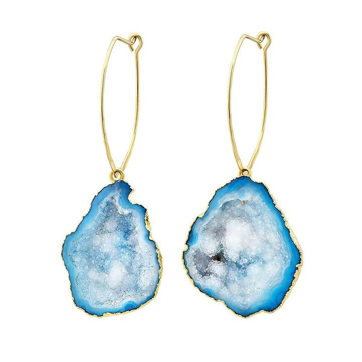 A pair of elegant Blue Nebula Earrings by Aria V featuring irregularly shaped blue geode stones with gold edges. Handmade in Ireland, the stones are attached to slim, elongated gold hoops, creating a striking and unique design.