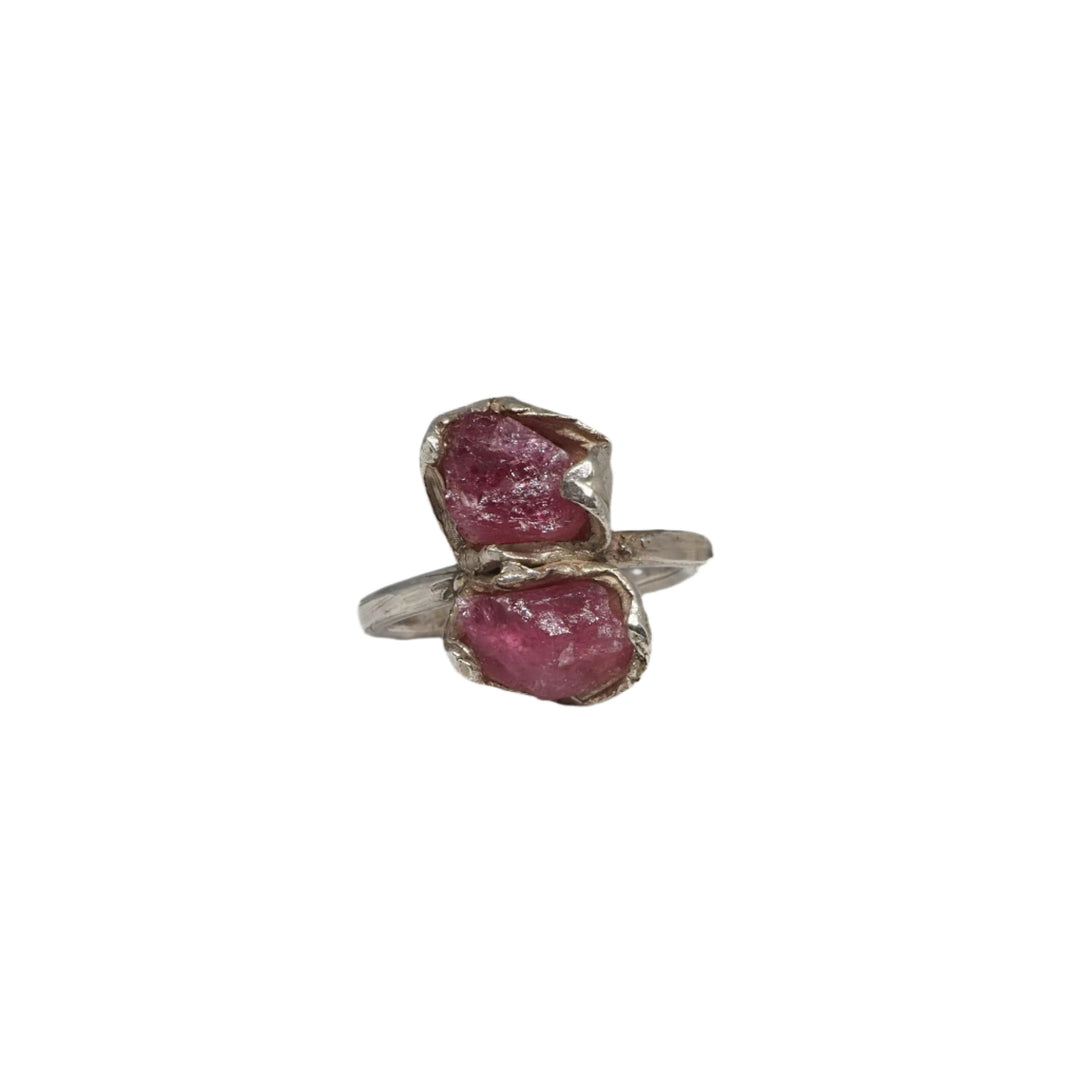 A Caroline Stokesberry-lee Double Ruby Ring featuring two raw pink gemstones set in a simple metal band is displayed on a textured white surface. In the background, there are large clusters of light blue and clear crystals. The overall aesthetic is natural and elegant.