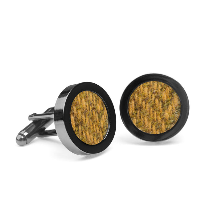 A pair of Amber Tweed Cufflinks by Orwell & Browne with round faces featuring textured, yellow-brown Donegal tweed enclosed in sleek black metal borders. The sustainable cufflinks are placed on a white surface, with one lying flat and the other standing upright.