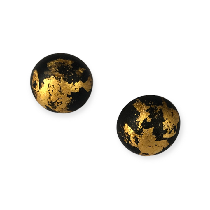 Two round black studs with irregular gold flecks on their surfaces, displayed against a white background. The Luxe Stud Earrings Black by Angela O'Keefe feature resin designs that give a marbled or cracked appearance, contrasting strikingly with the matte black base and sterling silver posts.