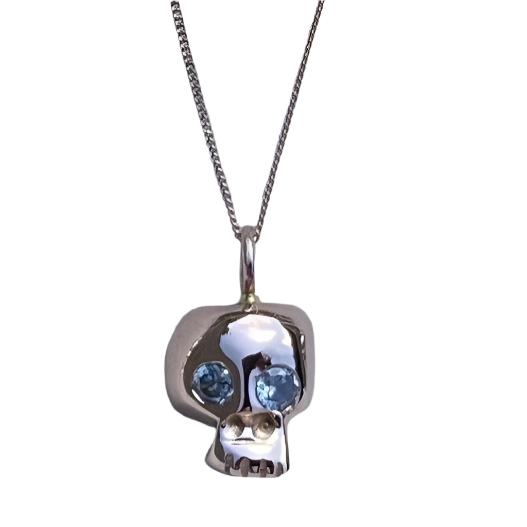 A 9ct Rose Gold Memento Mori Necklace from Cosmic Boulevard, featuring two blue gemstone eyes, is displayed on a light gray background. Handmade jewellery from a Dublin studio, the skull pendant has a smooth, reflective surface, and the chain is thin and delicate.