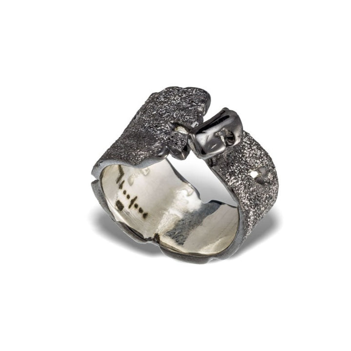 A silver ring with a rugged, textured design features an open, adjustable band that overlaps at the top. The inner side of the band is smooth and polished. Enhanced with Black Rhodium, the Rythmos Black Rhodium Ring by Theodora D exudes an organic, handcrafted appearance.