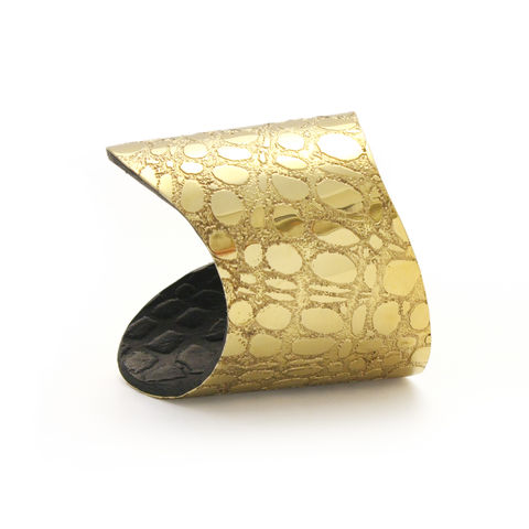 A close-up image of an elegant, wide cuff bracelet crafted from 18-carat gold with a textured pattern resembling scales or abstract shapes. The handmade bracelet has a slightly curved, open-ended design, exhibiting a shiny, metallic finish with a black inner surface. This is the Skin Textured Gold & Black Etched Cuff - Wide by Sara Gunn.