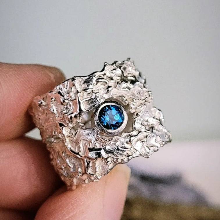 A person holds a uniquely textured "That Place Beyond  Wide Silver Ring with Gemstone" by Caroline Stokesberry-lee, part of the 2022 Collection, adorned with a dark blue gemstone at its center. The wide ring has an irregular, rugged design that gives it an organic appearance. The background is out of focus, emphasizing the intricate details of the ring.