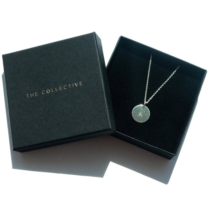 A Personalised Large Disc Charm in Silver is displayed inside an open black box. Hand stamped and elegant, this personalized gift is perfect for any occasion. The lid of the box, placed beside it, features "Personalised Charms" in gold lettering. The display is illuminated by natural light.