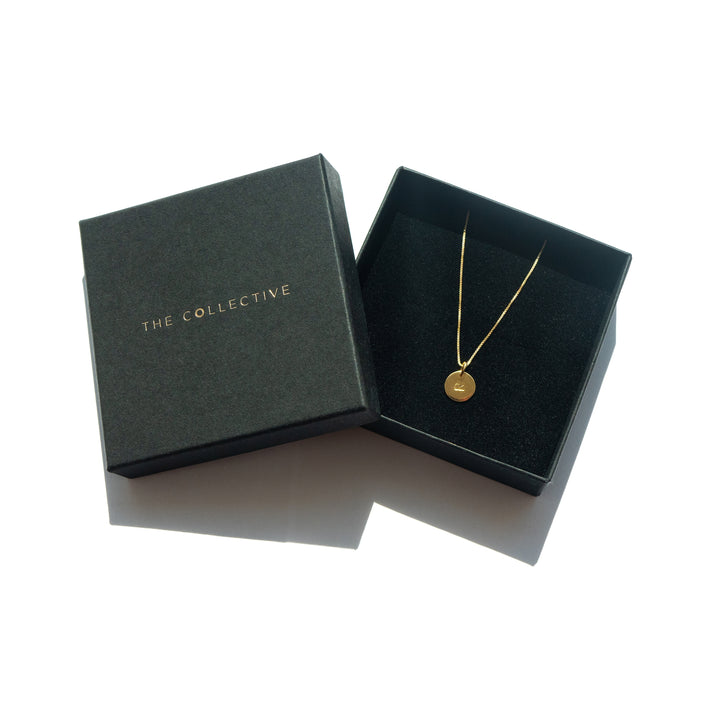 A black box with gold foil text reading "Personalised Charms" sits open, revealing a delicate gold necklace with a round, hand-stamped charm inside. The pendant features an engraved design and hangs from an adjustable chain, crafted from gold-plated silver. The box and necklace are set against a plain white background.