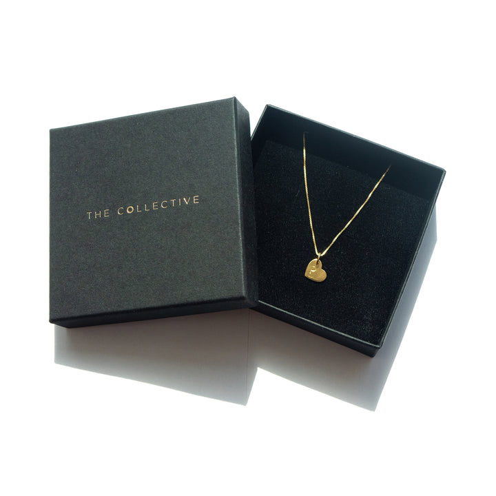 A Personalised Small Heart Charm in Gold by Personalised Charms is displayed in a black box with a matching lid. The lid, placed to the left, features the words "The Collective" in gold letters. The interior of the box is lined with soft black fabric and includes an adjustable oval belcher chain.