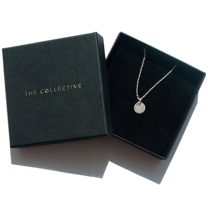 An open black jewelry box with "Personalised Charms" written in gold on the lid reveals a delicate silver necklace with a small round pendant. The Personalised Small Disc Charm in Silver appears to be hand stamped and shiny, resting against the black velvet interior of the box. The elegant piece hangs from an oval belcher chain.