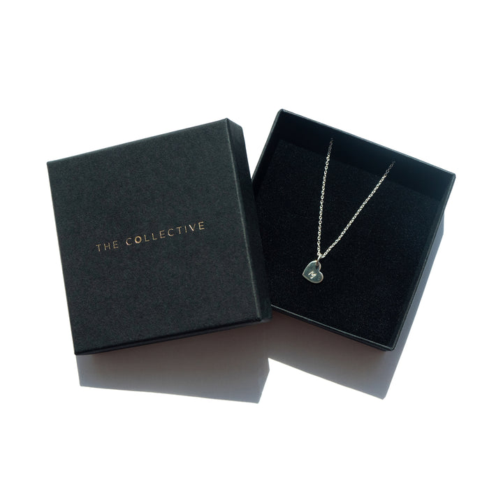 A Personalised Small Heart Charm in Silver with a small heart-shaped pendant featuring a tiny cross and silver charms is displayed in a black velvet-lined gift box. The oval belcher chain secures with a lobster claw fastening. The box lid reads "PERSONALISED CHARMS" in gold lettering, set against a plain white background.