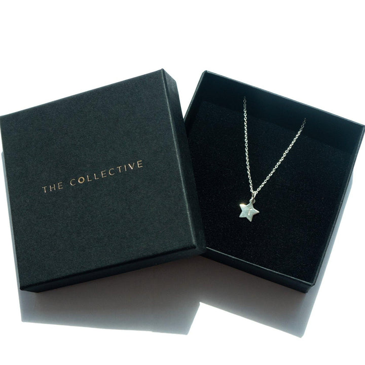 A Personalised Small Star Charm in Silver necklace, adorned with a charm and hung on an elegant oval belcher chain, lies in a black jewelry box. The matching lid is placed beside it, displaying "Personalised Charms" in gold letters. Both the box and the necklace are bathed in soft, natural light.