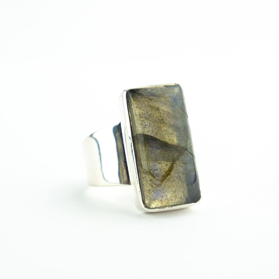 A Watermelon Tropical Silver Rectangle Labradorite Ring is displayed against a white background. The stone exhibits an iridescent mix of gold and blue hues, and the ring has a wide band.