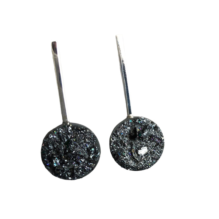 A pair of dark, glittery Urban Geode Silver Sphere Drop Earrings with silver hooks from Angela O'Keefe, set against a light gray background. The stones are round with a textured, sparkly surface, adding a touch of iridescence to the design.