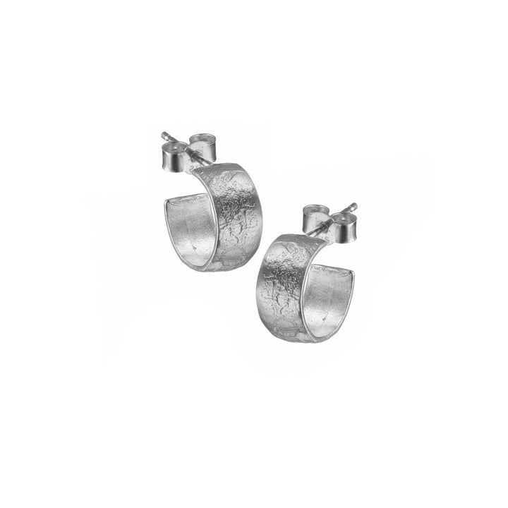 A pair of Lynsey de Burca Muir Sleepers with a textured, slightly hammered design. The earrings have a smooth, curved shape with posts and butterfly backings for secure fastening. The plain white background highlights the handmade jewellery details of the Muir Sleepers.