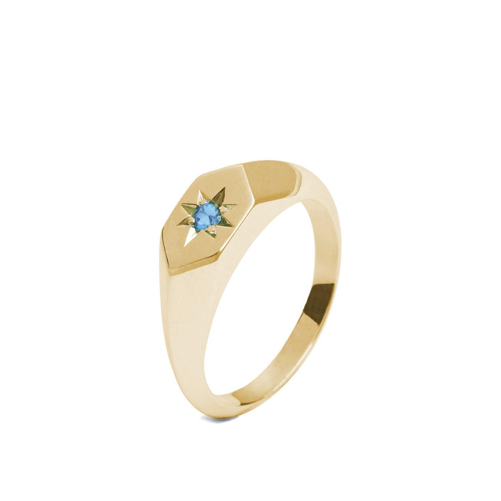 A **Cosmic Boulevard Starlight Aquamarine Birthstone 9ct Gold Signet Ring** featuring a hexagonal face with a star-shaped design. At the center of the star is the birthstone of March, a small blue gemstone. The band is sleek and polished, complementing the geometric and elegant design of the ring.