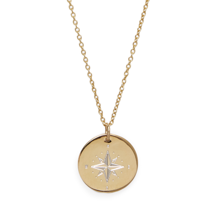 A Compass Symbol Pendant by The Collective Dublin featuring a circular pendant adorned with an engraved compass rose design. The pendant is polished and slightly reflective, resembling a sterling silver engraved disc, and it hangs from a delicate, gold chain.