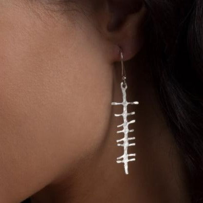 Close-up of a person's ear wearing a sterling silver earring shaped like a fish skeleton. The earring hangs and catches the light against the person's skin and dark hair, reminiscent of Ogham Treasure Love (Grá) Rustic Earrings.