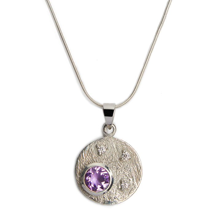 The Gallardo & Blaine Designs Celeste Necklace in Silver & various gemstones is crafted from sterling silver, featuring a round pendant with a textured surface adorned with small cubic zirconia stones and a larger central purple gemstone. The thin, smooth chain accentuates the elegance of this exquisite piece.