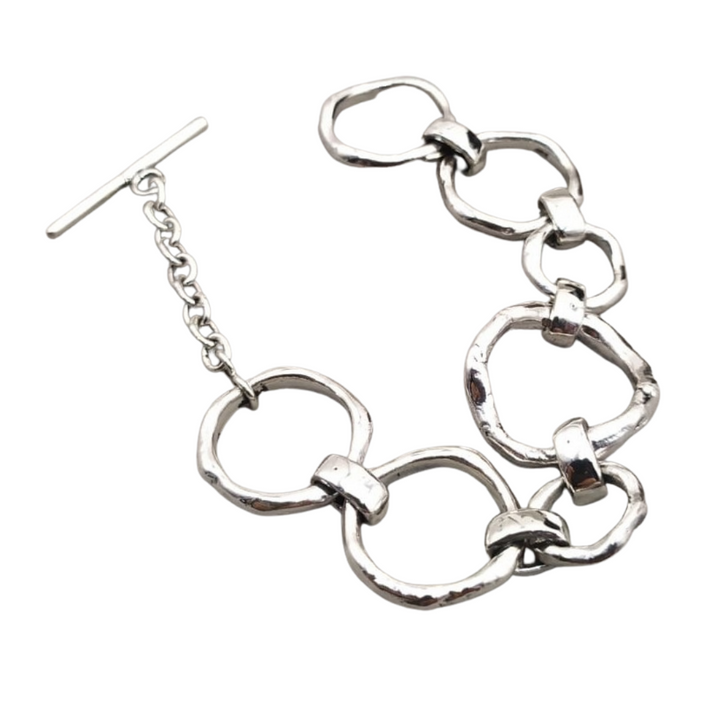 A stylish Sea Honey Bracelet Silver made of linked, thick silver rings is laid flat on a white background. It features a T-bar clasp with an adjustable chain, giving it a modern and elegant appearance. The metal has a slightly hammered texture, adding a touch of uniqueness to the new design by Gallardo & Blaine Designs.