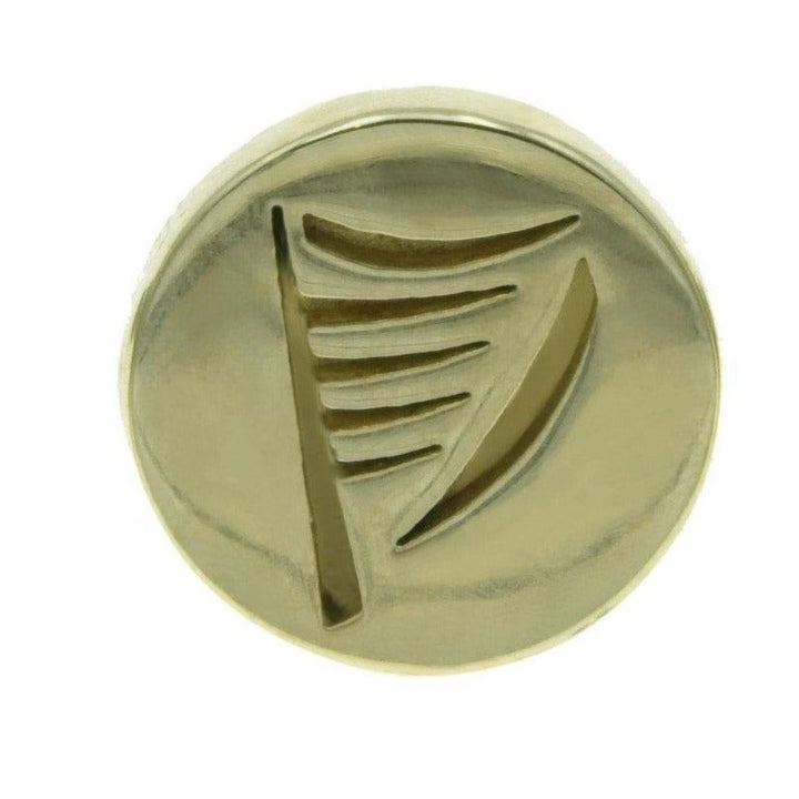 A round, gold-colored *Harp Lapel Pin* by *Millett Wade* features a design with five parallel lines converging to a central point, resembling a stylized harp or musical symbol. Reminiscent of hand-finished brass items, the pin surface is glossy and has a smooth finish.