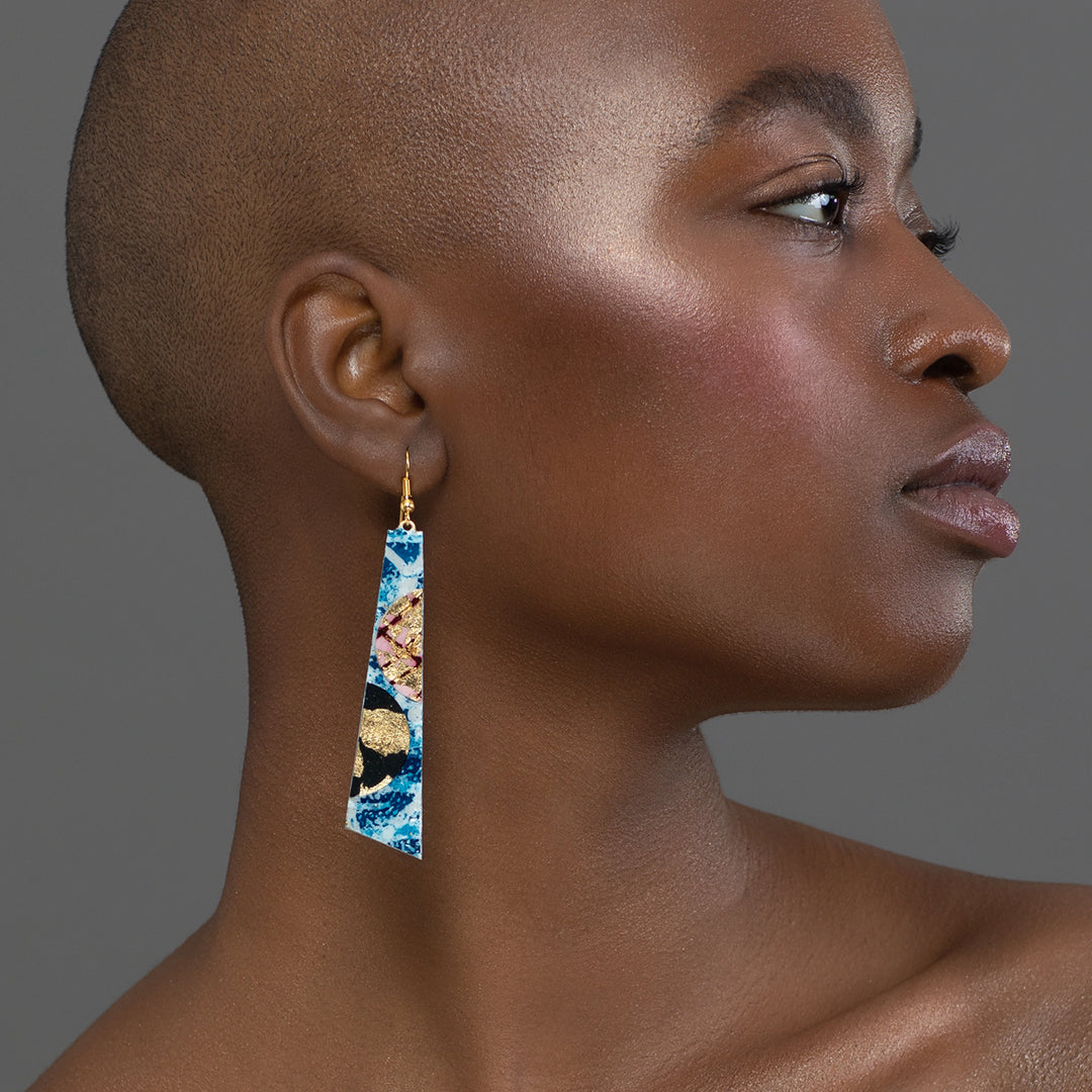 A person with a shaved head is shown in profile view against a gray background. They are wearing the Rothlú Hoop-La Batik Textile Earrings, which are vibrant and triangular with hues of blue, red, black, and gold featuring unique texture. The individual has clear skin and is looking straight ahead.