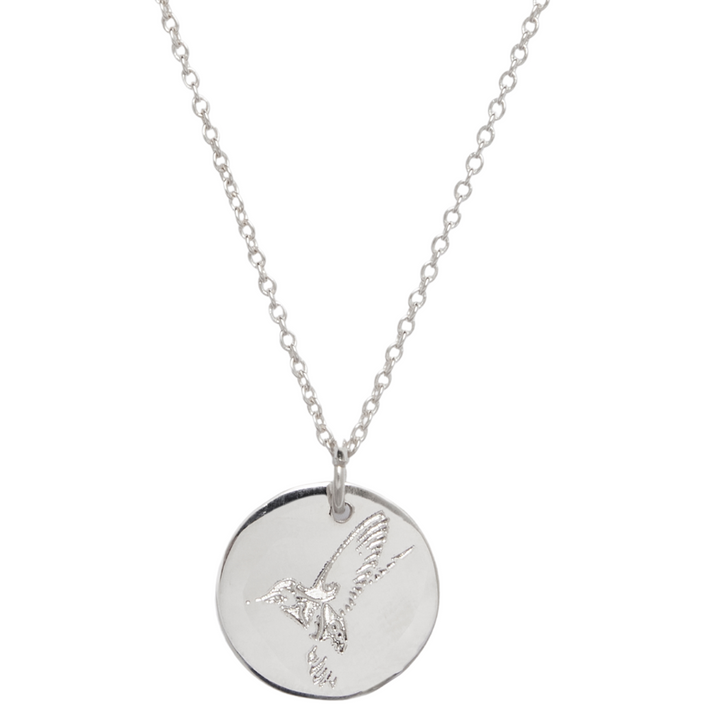 The Collective Dublin's Hummingbird Symbol Pendant features a sterling silver necklace with an engraved disc pendant showcasing a hummingbird design. The pendant hangs from a delicate chain, creating an overall look that is both elegant and minimalistic.