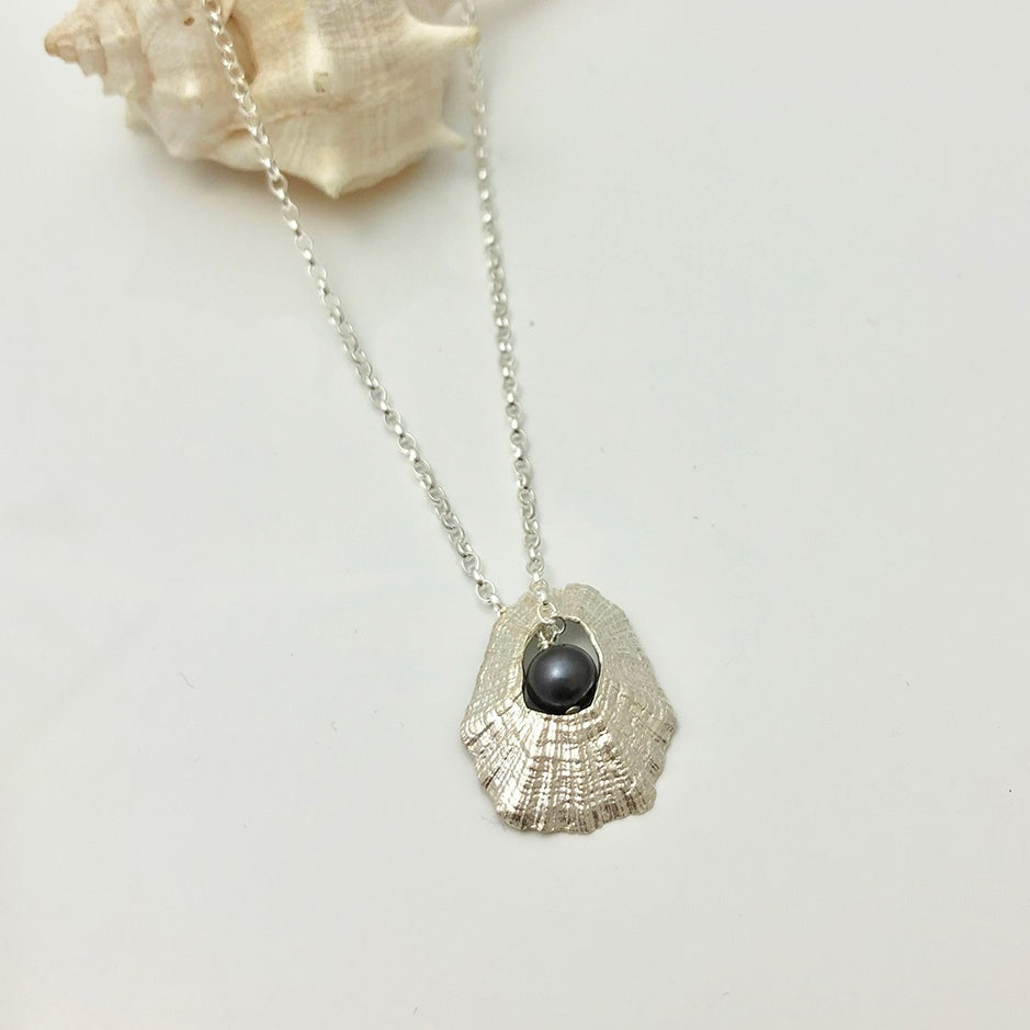 The Hollow Limpet Shell Necklace with Pearl Small by Jennifer Kinnear features a sterling silver textured shell pendant with a freshwater pearl at the center, displayed on a light background with a seashell in the corner.