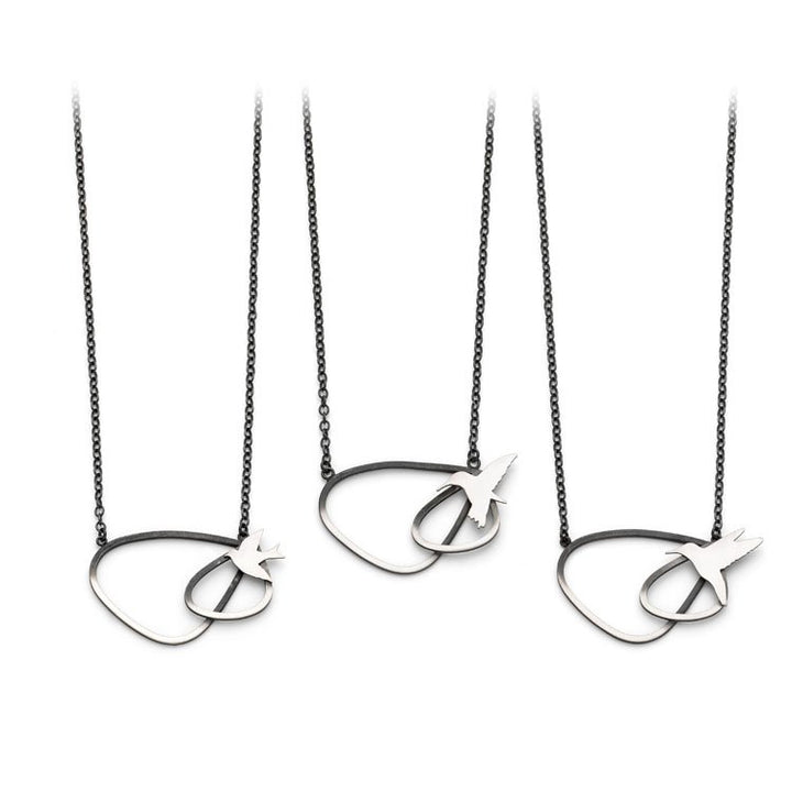 Three identical necklaces are displayed, each featuring a black chain and a distinctive pendant. The looped design with a small geometric arrowhead shape is crafted from oxidised silver-colored metal. The symmetrical arrangement showcases the Leoma Drew Two Tone Silver Wing Pendant Necklace with Birds evenly spaced, resembling elegant bird designs in flight.