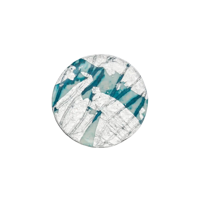 A hypoallergenic silver-plated Ró Beag Dress Ring in Silver/Sea Blue by Rothlú lies in an open gray box with black lining. The bottom half of the box lid is visible, displaying a partial logo in teal. Green leaves rest beside the box on a white surface.