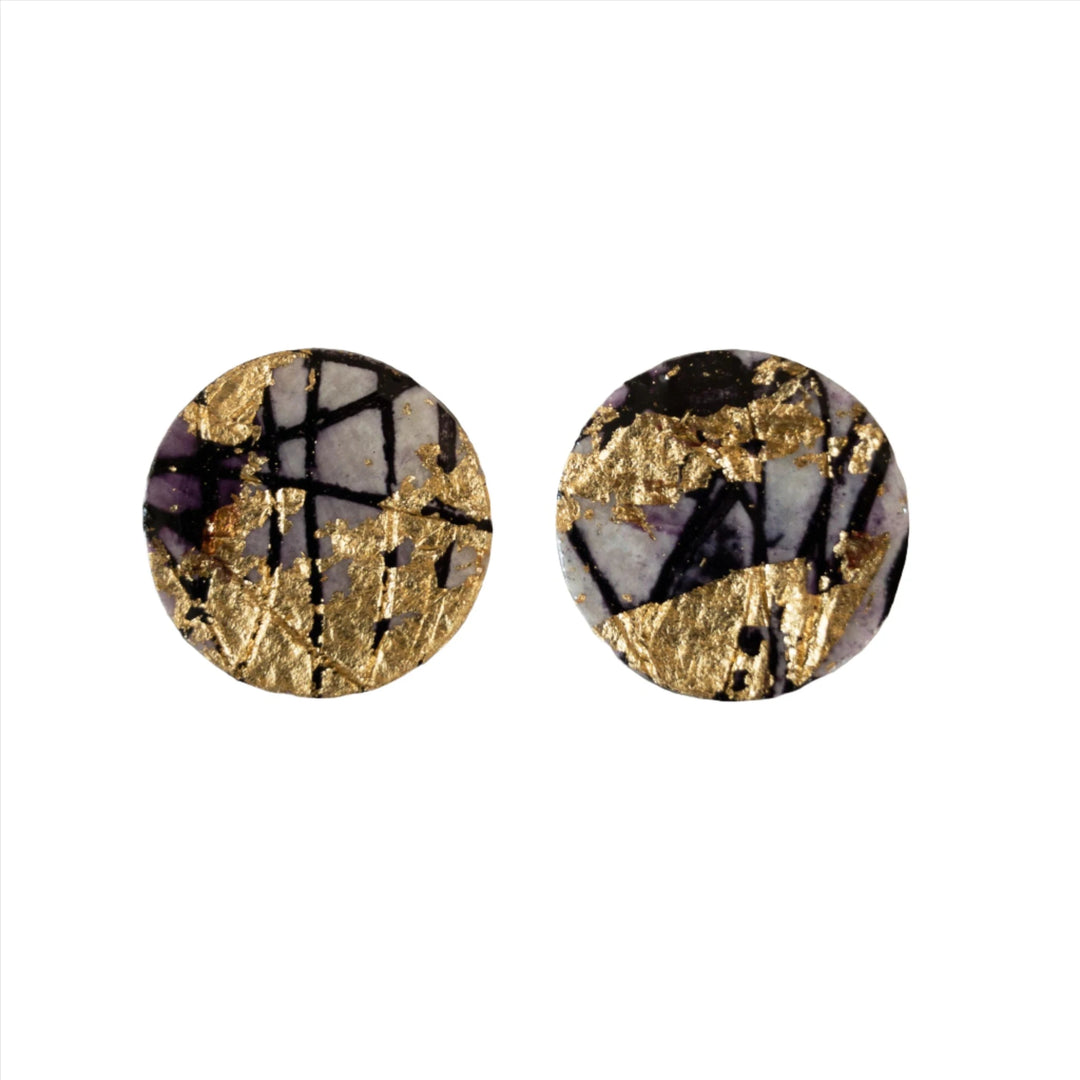 Round sgraffito batik earrings with a design resembling black, white, and purple stone, accented by gold leaf patterns. These one-of-a-kind Ró Sgraffito Stud Earrings in Gold/Black by Rothlú rest on a smooth, white surface, showcasing their artistic and elegant appearance. They feature sterling silver posts for comfort and durability.