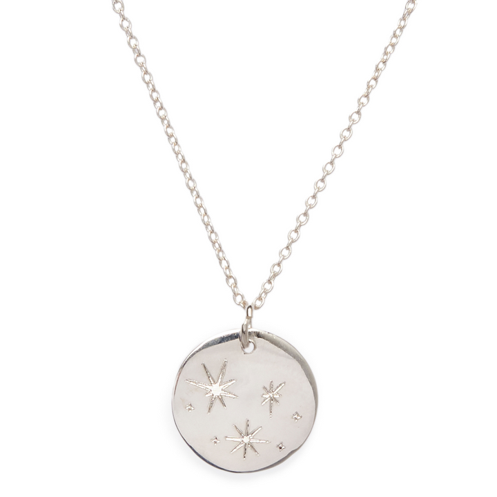 A silver necklace features a round engraved disc pendant with a design of stars and small dots. The Stars Symbol Pendant from The Collective Dublin hangs from a delicate chain, giving it a minimalist and elegant appearance, perfect for those who love personalized jewelry.