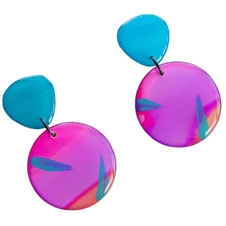 The Sunset Surf Statement Earrings by Naoi are a pair of vibrant, colorful earrings with teal-blue pebble-shaped tops and large, round pink-purple bottoms featuring abstract teal accents. These hand-painted earrings have a glossy finish and a modern, artistic design, attached to durable stainless steel earring posts for lasting wear.