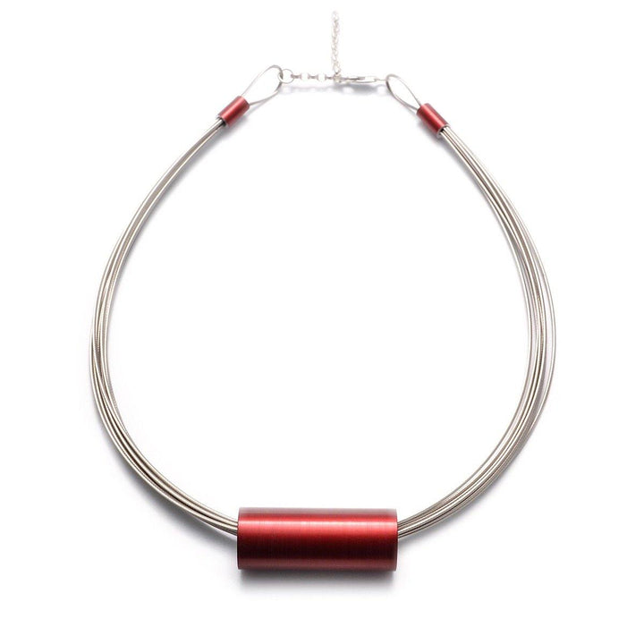 Introducing the Tensor Tube Pendant by Filip Vanas: A minimalist necklace featuring three strands of thin, metallic cables gathered together and accented with a cylindrical, red bead at the center. Crafted from stainless steel necklets, the ends are secured with small red connector tubes and a silver clasp.