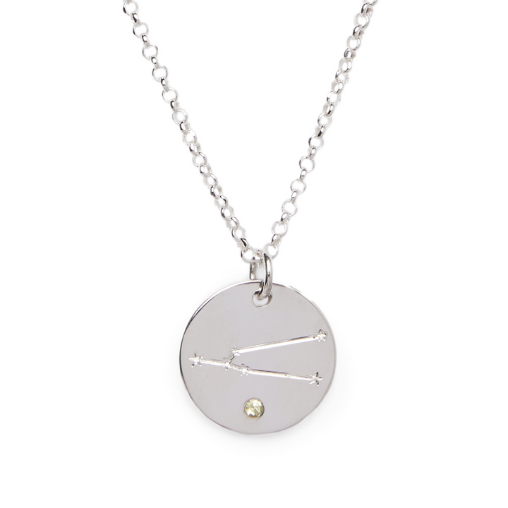 A silver necklace features a round, engraved disc pendant showcasing a constellation and a small birthstone at the bottom. The Constellation Symbol Pendant by The Collective Dublin hangs from a delicate chain with evenly spaced links.