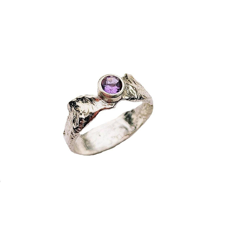 This Faerie Tale Ring Thin Band by Caroline Stokesberry-lee showcases a textured silver band with a rough, organic design and a bezel-set round purple gemstone. The uneven, natural finish lends it a rustic charm, while the gem adds a pop of color to the silver tone.