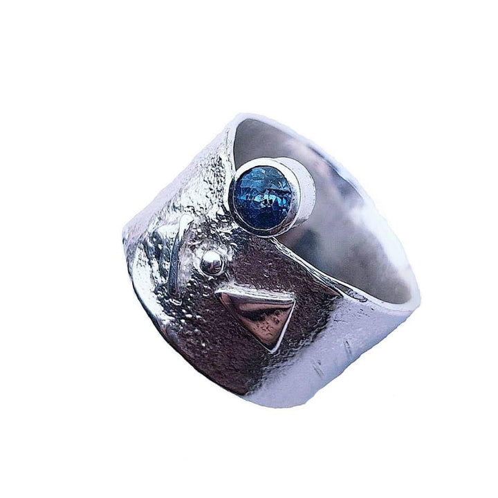 A wide sterling silver Blue Topaz Guardian Ring by Caroline Stokesberry-lee features a hammered texture and geometric designs, including a triangular shape and a small sphere. The focus is a blue topaz gemstone set in a raised bezel, adding a pop of color to the metallic surface.