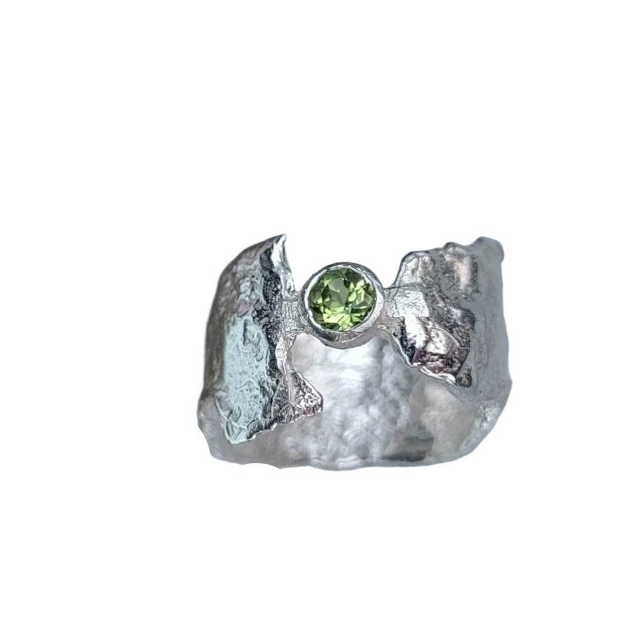 A handcrafted sterling silver ring featuring a textured band and a shimmering peridot gemstone at its center is displayed on a white background with text. Delicate flowers and greenery contribute to the faerie tale aesthetic of this enchanting piece of jewelry: the Peridot Faerie Tale Ring by Caroline Stokesberry-Lee.