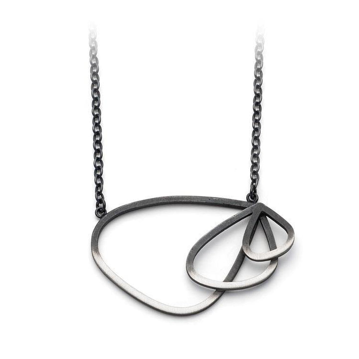 A necklace with a minimalist design, featuring a Two Tone Triple Wing Pendant by Leoma Drew. The pendant, crafted from dark metal, hangs from a delicate trace chain. The larger teardrop has an asymmetrical shape, while the smaller teardrop is nested inside it.