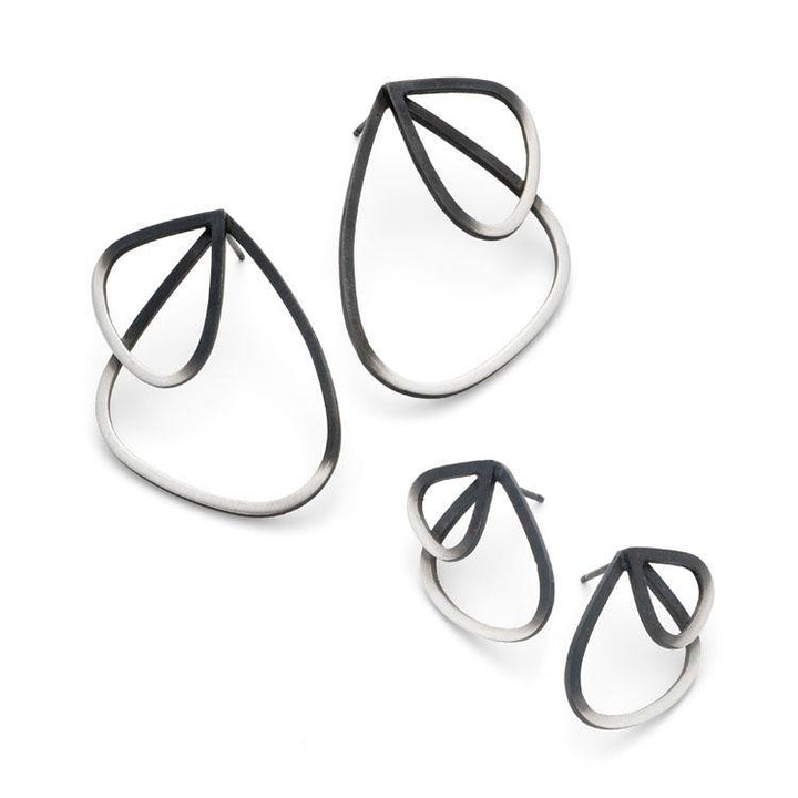 A pair of large black and oxidized sterling silver teardrop-shaped hoop earrings and a matching pair of smaller teardrop-shaped stud earrings, known as the Two Tone Double Wing Studs by Leoma Drew, are displayed on a white background. The design, available in various sizes, features overlapping metal loops with a modern and artistic style.